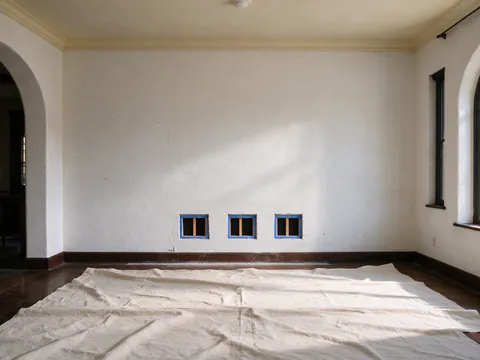A small square opening cut neatly in a plaster wall above a baseboard in a Los Angeles Craftsman, wood lath visible behind it and a drop cloth on the floor